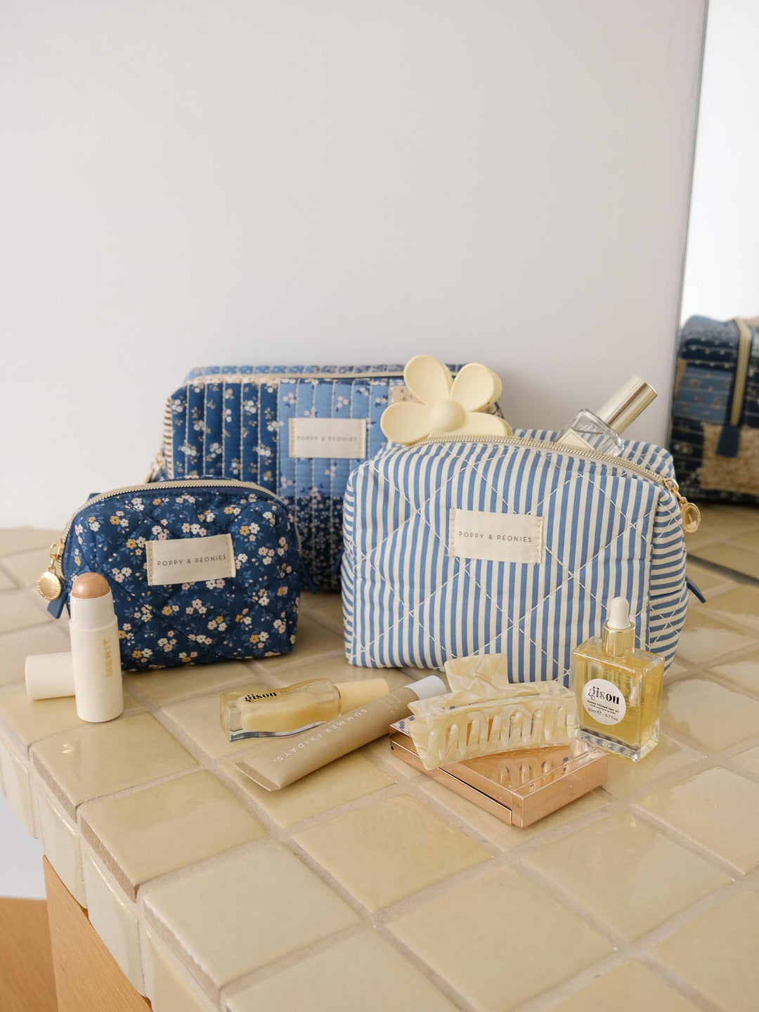 Product-focused lifestyle shot of the Cosmetic Trio in Navy Patchwork, featuring all three coordinating cosmetic cases in blue floral, striped, and patchwork prints styled alongside beauty essentials.