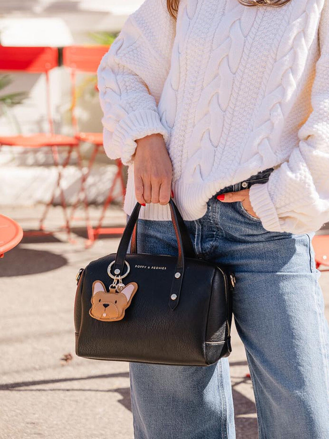 Person holding a black handbag with a dog bag charm, wearing a white sweater and blue jeans.