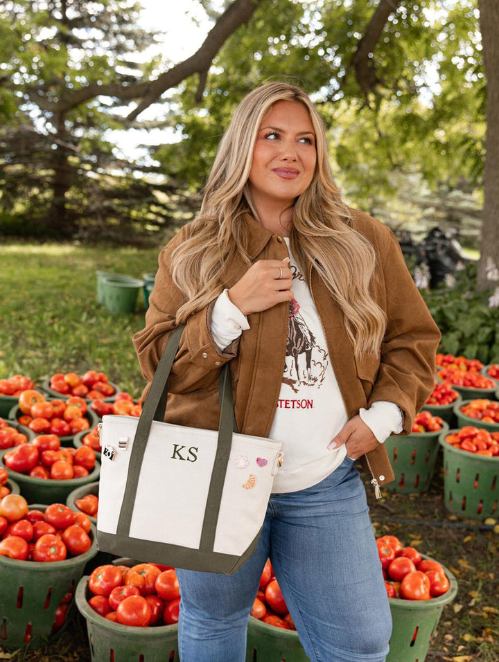 Poppy & Peonies The Small Signature Tote in Olive carried by its olive green handles, styled with a brown jacket and denim jeans at an tomato orchard.
