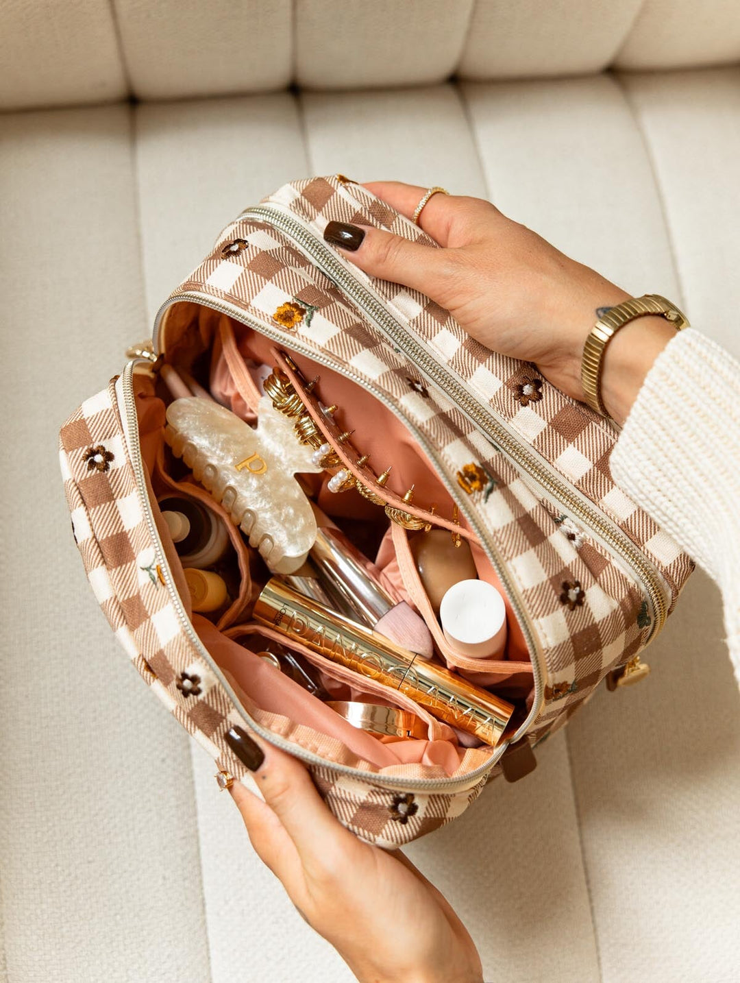 Poppy & Peonies Double Take Cosmetic Case in October Gingham, shown open on a cream chair with brown and cream gingham fabric, floral details, blush pink lining, and makeup essentials organized inside.