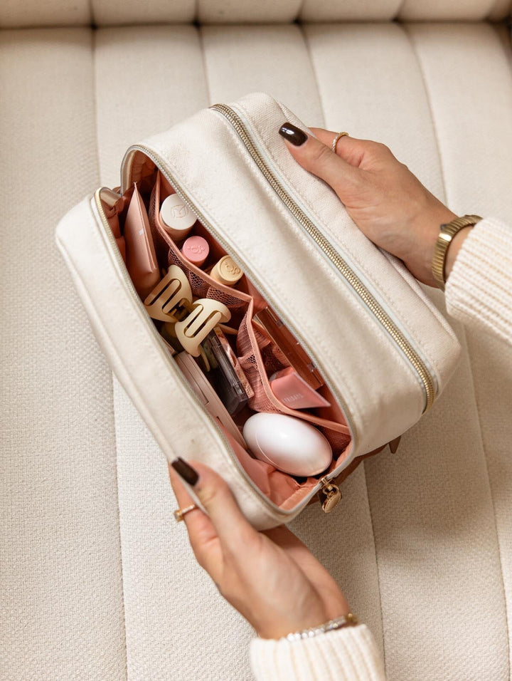 Poppy & Peonies The Double Take Cosmetic in Olive held open to reveal two zippered compartments filled with makeup products and skincare essentials.