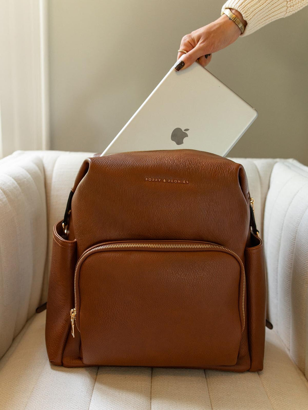 Poppy & Peonies The Cabin Backpack in Chestnut resting on a cream chair as a hand places a silver laptop into the main compartment.