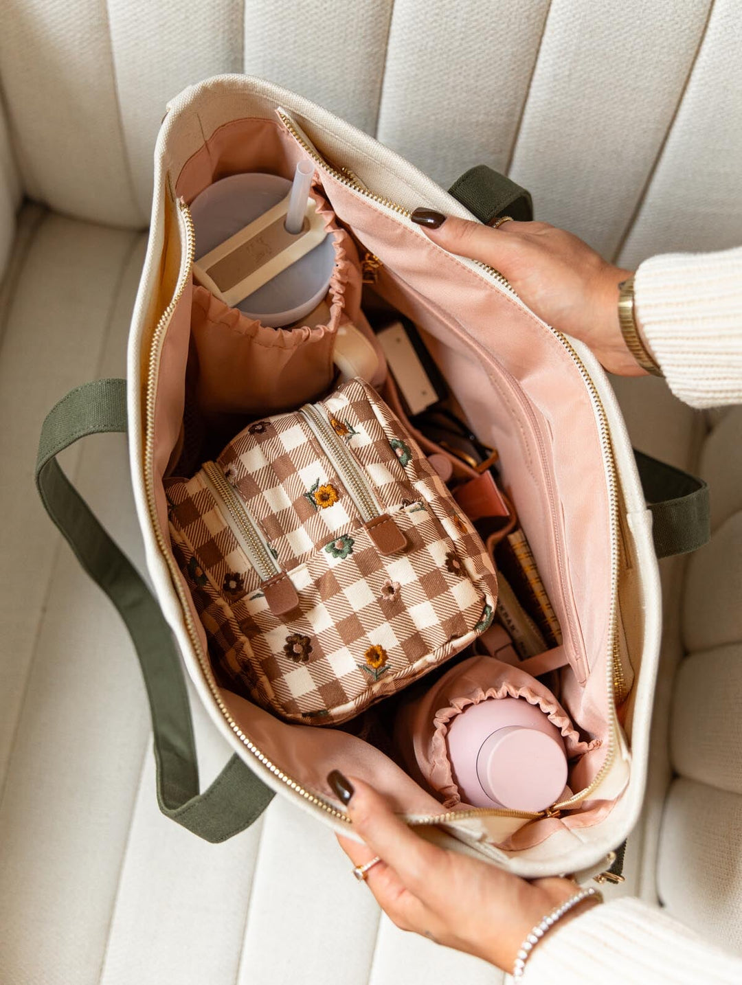 Poppy & Peonies The Signature Tote in Olive, shown open on a cream chair with blush pink lining, olive green handles, interior organizer pockets, and a Double Take Cosmetic Case in October Gingham tucked inside alongside water bottles and everyday essentials.
