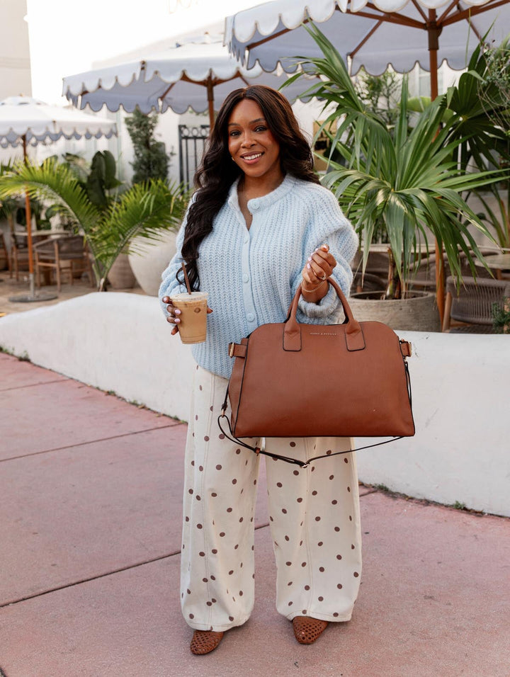 Woman holding a brown handbag and a coffee cup outdoors with plants and tables in the background