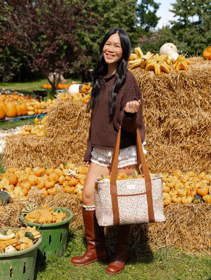 Poppy & Peonies Mini Traveller in October Floral, held by a woman at a pumpkin patch, highlighting its roomy rectangular silhouette, cream floral canvas exterior, and long brown shoulder straps.