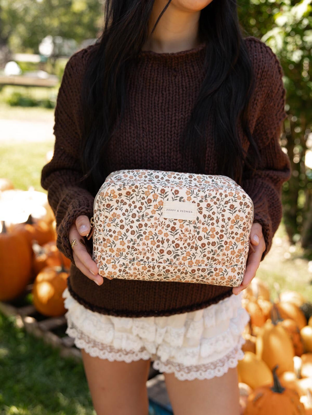 Poppy & Peonies Double Take Cosmetic Case in October Floral, held by a woman wearing a brown sweater and cream shorts outdoors near a pumpkin display, showcasing its cream floral-printed exterior.