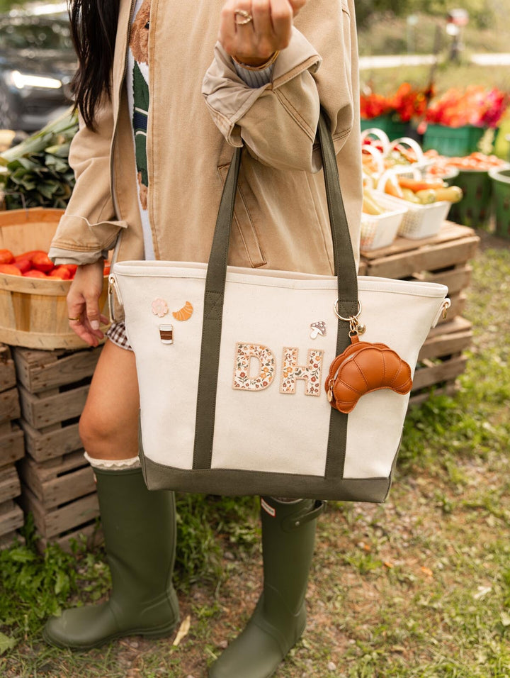 Poppy & Peonies floral letter patches and decorative signature pins on the Signature Tote in Olive, carried by a woman wearing green boots at market. 