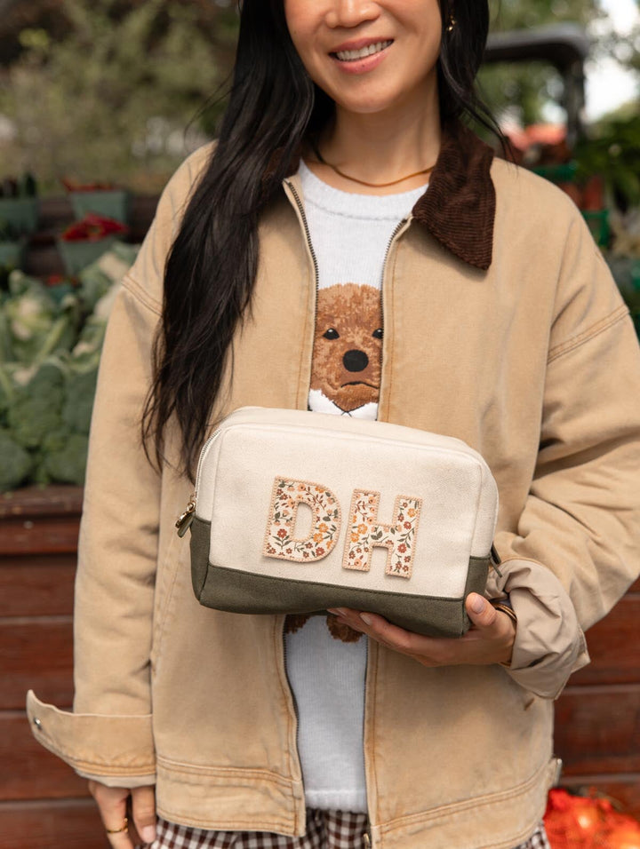 Poppy & Peonies The Double Take Cosmetic in Olive personalized with decorative letter patches, held by a woman wearing a tan jacket at an outdoor market.