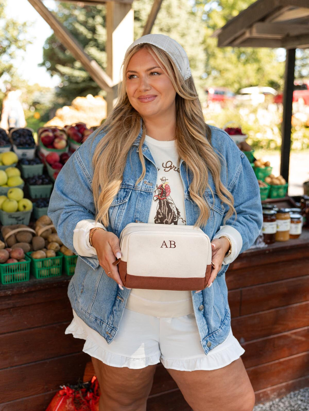 Poppy & Peonies The Double Take Cosmetic in Chestnut featuring AB initials on the cream exterior, held by a woman standing in front of a colorful produce stand.