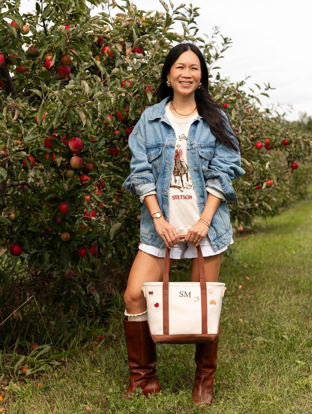 Front lifestyle view of Poppy & Peonies The Small Signature Tote in Chestnut, held by a woman standing in an orchard wearing a denim jacket and brown boots.