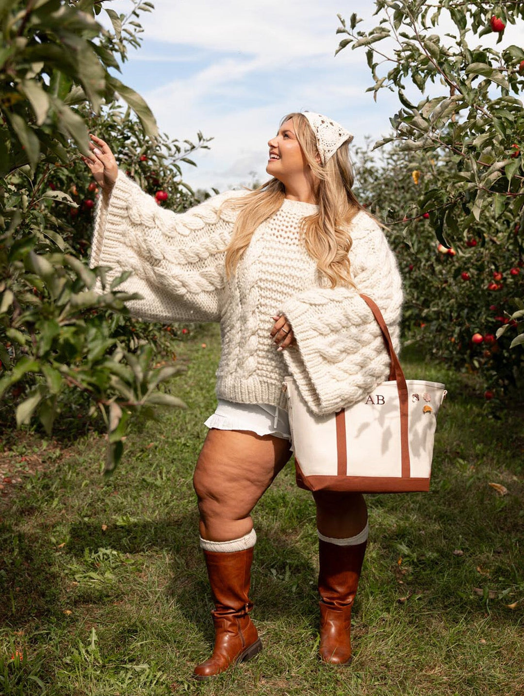 Woman reaching toward an apple tree while carrying Poppy & Peonies The Signature Tote in Chestnut by its long brown handles.