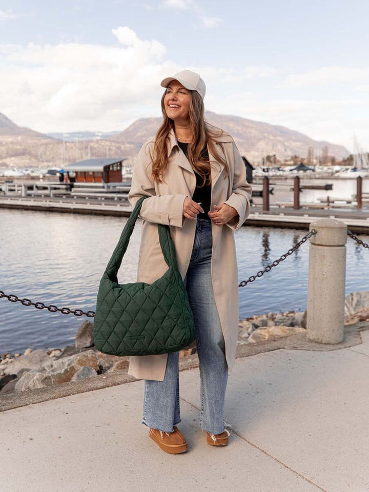 Woman with a green quilted hobo bag standing by a waterfront with mountains in the background