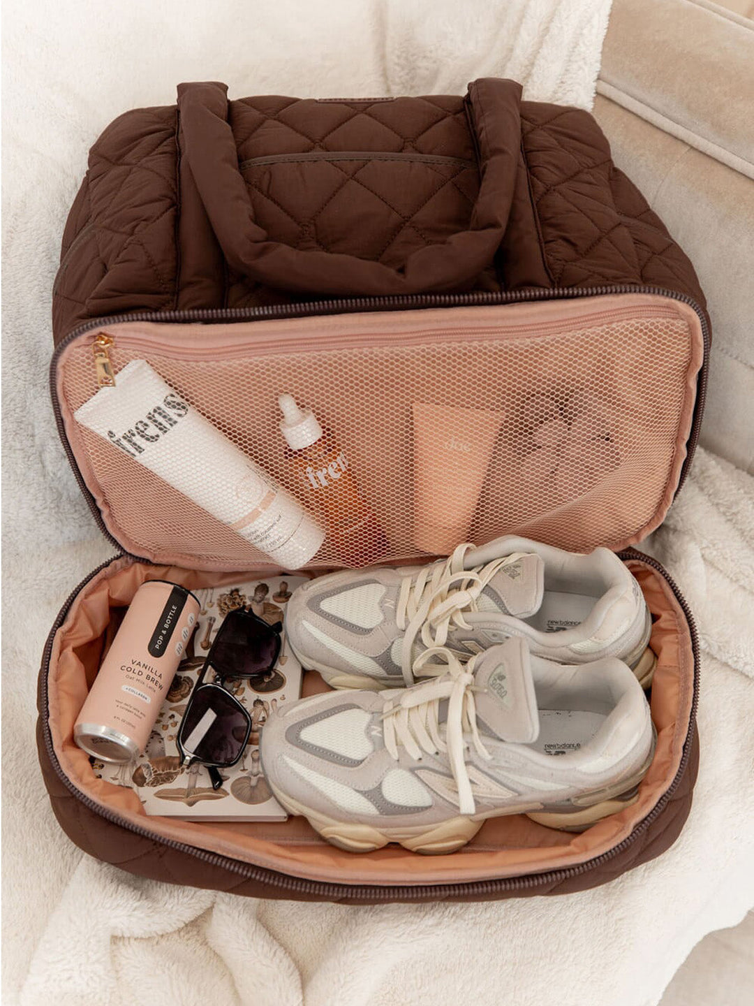 Brown travel bag with pink interior containing sneakers, skincare products, and sunglasses.